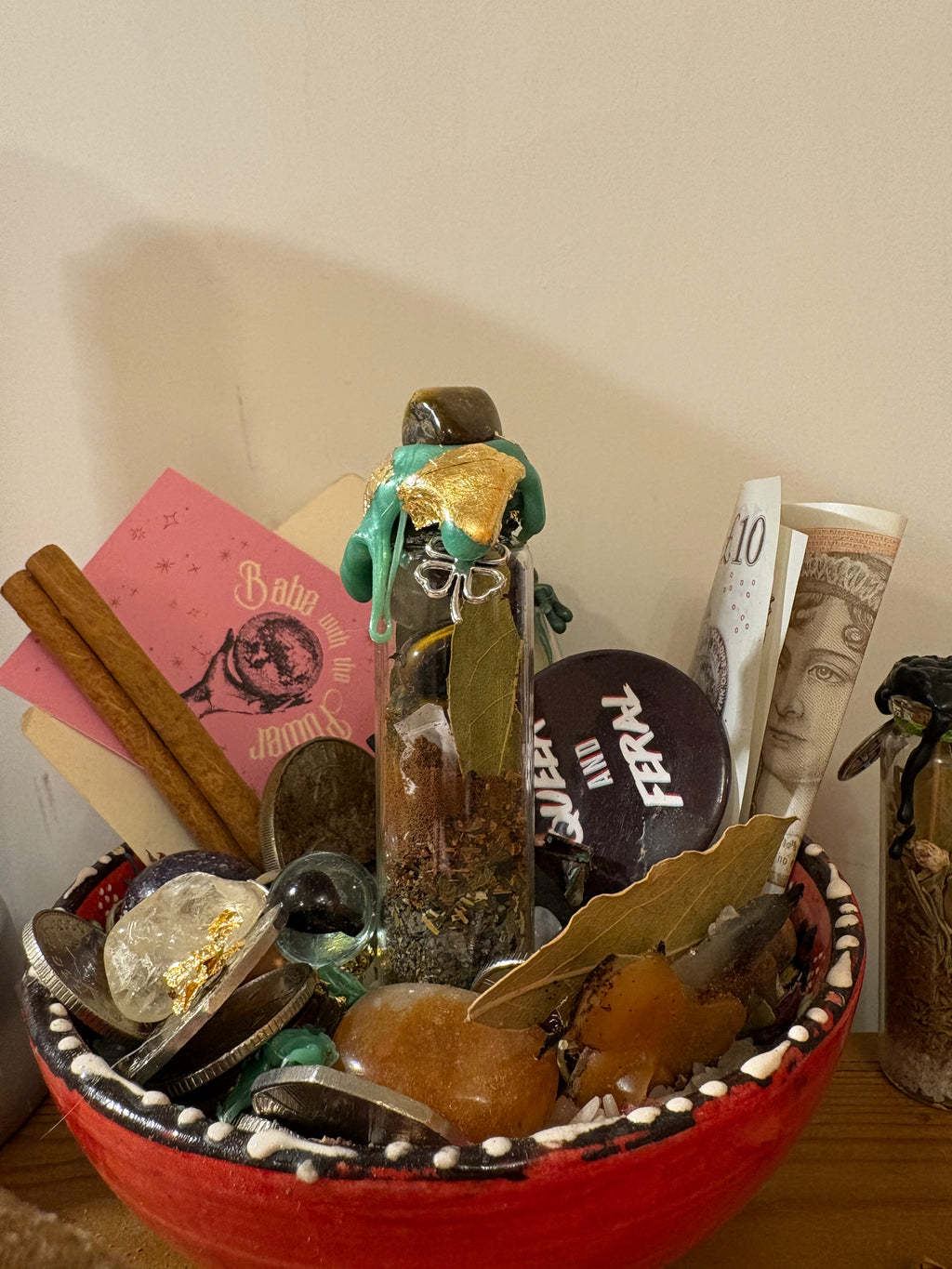 A decorative ritual or altar bowl filled with crystals, herbs, coins, a small glass vial with a figurine, and rolled paper, photographed indoors against a plain wall.